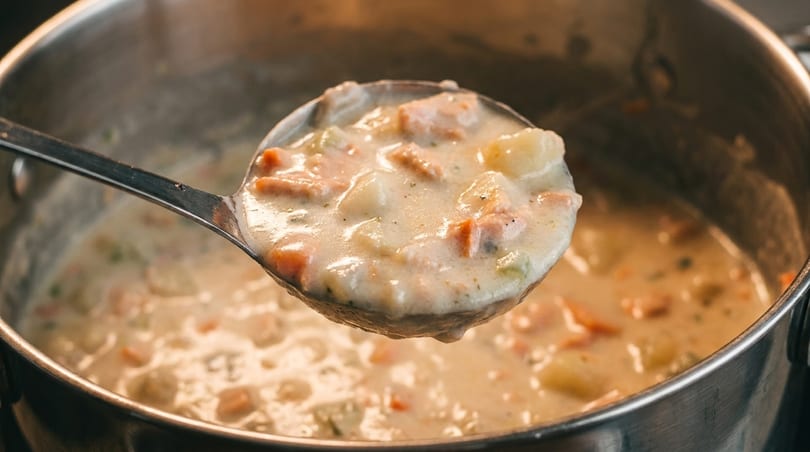 A ladle full of creamy clam chowder with potatoes, carrots, and celery, lifted from a pot.