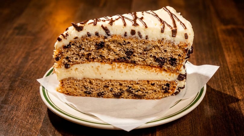A slice of layered chocolate chip cake with white frosting and chocolate drizzle on a white plate.